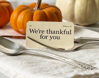May include: A small card with the text "We're thankful for you" on a white linen surface with two orange pumpkins and a white pumpkin. The card is resting on a silver spoon.