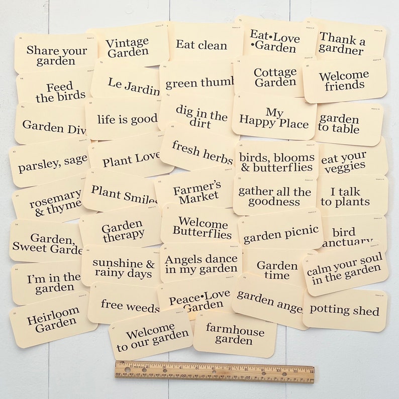 Pu&ograve; includere: Una collezione di carte color crema con testo nero, ciascuna con una frase diversa sul tema del giardinaggio. Le carte sono sparse su una superficie di legno con un righello nell'angolo in basso a destra.