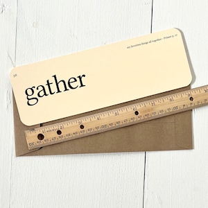 May include: A cream-colored card with the word "gather" printed in black on the front. The back of the card has the text "my favorites things all together - Primer p. 17" printed in black. The card is lying on a brown envelope.