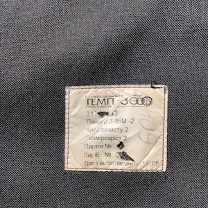 Op de afbeelding: Close-up van een zwarte, getextureerde stof met een rechthoekig label. Het label is gebroken wit met zwarte tekst, inclusief cijfers en Cyrillische tekens. Het label lijkt een productlabel te zijn.