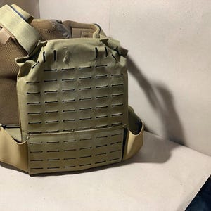 Op de afbeelding: Tan tactische plate carrier vest met meerdere rijen horizontale sleuven voor het bevestigen van zakjes en uitrusting. De vest heeft verstelbare schouderbanden en een cummerbund. Het materiaal lijkt een duurzame, heavy-duty stof te zijn.