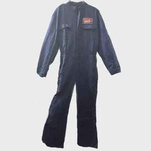 Op de afbeelding: Marineblau jumpsuit met lange mouwen, een kraag en meerdere zakken. Een rechthoekige patch met rode en witte tekst staat op de borst. De jumpsuit heeft een riem in de taille en wijde pijpen.