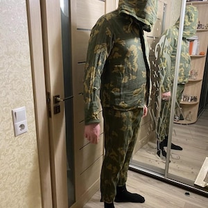 Op de afbeelding: Een camouflagejas met capuchon en bijpassende broek in groen- en bruintinten. De outfit wordt gedragen met zwarte sokken. De persoon staat voor een spiegel.