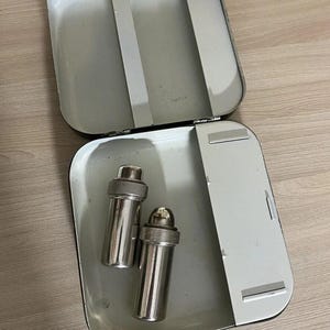 Op de afbeelding: Een open, rechthoekige, witte metalen container met twee zilveren cocktailshakers erin. De container heeft een scharnierend deksel en een handvat. De shakers zijn cilindrisch met een afgeronde bovenkant. De achtergrond is een lichtbruin houten oppervlak.