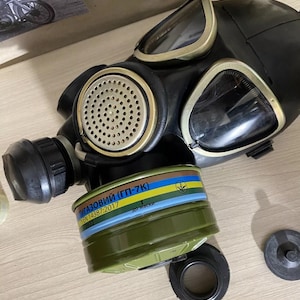 Puede incluir: Máscara de gas negra con lentes transparentes y un filtro circular. El filtro es verde oliva con rayas azules, amarillas y verdes. La máscara tiene un altavoz redondo perforado y una conexión roscada para el filtro.