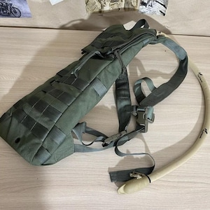 Op de afbeelding: Een olijfgroene hydratatiepack met meerdere bevestigingspunten en verstelbare bandjes. De pack bevat een beige drinkslang en een bijtventiel. De pack is ontworpen voor buitenactiviteiten en tactisch gebruik.