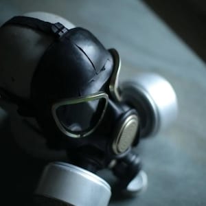Puede incluir: Una máscara de gas negra y plateada con una cubierta facial de goma negra y dos botes plateados. La máscara tiene una cubierta para la cabeza blanca y está sobre un fondo gris.