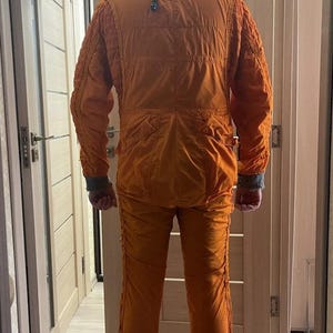 Op de afbeelding: Een oranje vliegpak met bijpassende helm. Het pak heeft meerdere zakken en geribbelde details op de schouders en mouwen. De helm is bruin met een donker vizier en een kinband. De persoon staat in een deuropening.