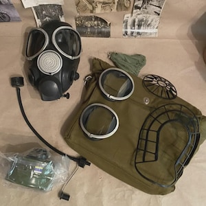 Puede incluir: Una máscara de gas negra con lentes transparentes y una cubierta de filtro plateada se muestra junto a una bolsa de lona verde con lentes transparentes y un marco de alambre negro. También hay un filtro verde y un cartucho de filtro verde.