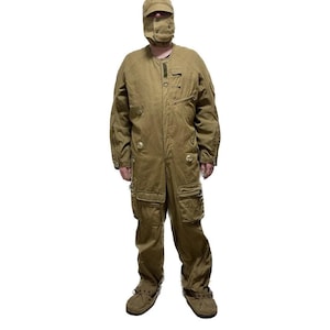 Op de afbeelding: Een bruinbeige overall met bijpassende hoed en gezichtsbedekking. Het eendelige kledingstuk heeft meerdere zakken, knoopsluitingen en een relaxte pasvorm. De outfit bevat bijpassende schoenen. De algehele stijl suggereert een werk- of utiliteitskledingstuk.