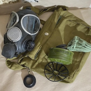 Puede incluir: Una máscara de gas negra con lentes transparentes, un filtro verde y una bolsa de lona verde oliva. La máscara tiene un filtro circular y una boquilla de goma negra. También se incluye una pieza circular negra y un marco de alambre negro.