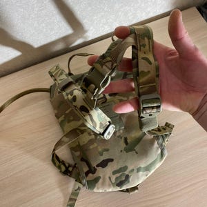 Op de afbeelding: Een camouflage hydratatiepack met verstelbare bandjes en plastic gespen. De pack heeft een gebogen ontwerp en een slangbevestigingspunt. Het kleurenschema omvat tinten groen, bruin en bruin, typisch voor militaire uitrusting.