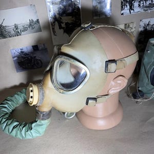 Könnte beinhalten: Eine Vintage-Gasmaske in Beige- und Olivgrüntönen, mit einem klaren Visier und einem flexiblen grünen Schlauch. Die Maske ist auf einem Schaufensterkopf montiert, mit Riemen und einem Filter. Historische Militärausrüstung.
