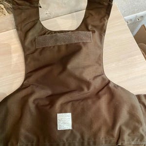 Op de afbeelding: Bruin kogelvrij vest met een nekopening en schouderstukken. Het vest heeft een rechthoekige patch met tekst en een klittenbandsluiting. Gemaakt van duurzaam, zwaar materiaal.