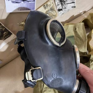 Puede incluir: Una máscara de gas negra con una lente circular y ribete plateado. La máscara tiene una textura engomada y está marcada con el número "1" y la fecha "2019". Equipo militar vintage.
