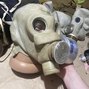Op de afbeelding: Een lichtgroen vintage gasmasker met ronde kijkglazen en een filterbus. Het masker heeft een rubberen gezichtsafdichting. Andere gasmaskers zijn zichtbaar op de achtergrond, wat wijst op een verzameling.
