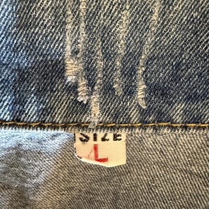 May include: Close-up of a denim garment, showing the fabric's texture and stitching. A tag with the word "SIZE" and the letter "L" in red is visible. The denim is a faded blue color.