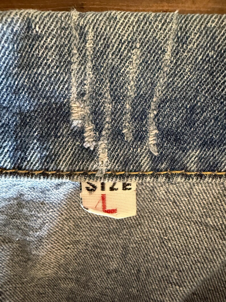 May include: Close-up of a denim garment, showing the fabric's texture and stitching. A tag with the word "SIZE" and the letter "L" in red is visible. The denim is a faded blue color.