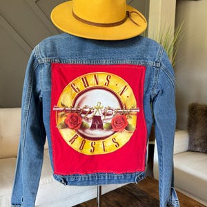 May include: A blue denim jacket with a Guns N' Roses logo patch on the back. The patch is red with a circular design featuring a skull, roses, and the band's name. A yellow hat with a brown band sits above the jacket.