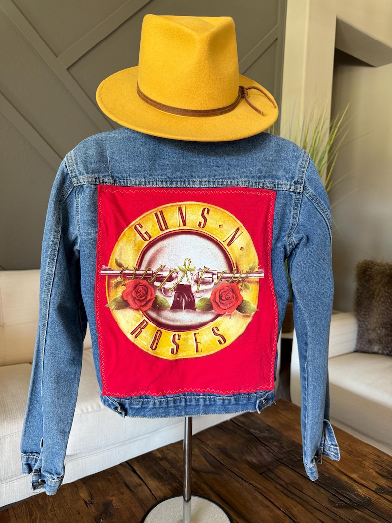 May include: A blue denim jacket with a Guns N' Roses logo patch on the back. The patch is red with a circular design featuring a skull, roses, and the band's name. A yellow hat with a brown band sits above the jacket.