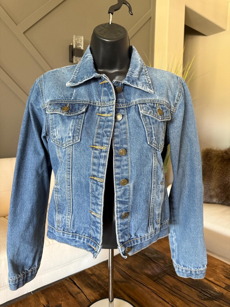May include: A light blue denim jacket with a classic design. The jacket features a collared neckline, button-down front, and two chest pockets. The jacket is displayed on a black mannequin.