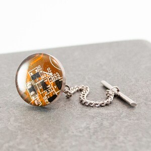 Op de afbeelding: Een zilveren stropdasgesp met een ronde, doorzichtige harsdop met een bedrukt printplaatontwerp in oranje en bruine tinten.