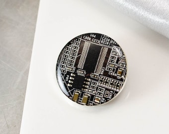 May include: A round, black and silver pin with a circuit board design. The circuit board has text on it, including "U44", "C609", "C273", "R259", and "C490".