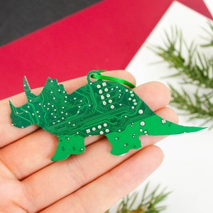 Könnte beinhalten: Ein grüner, dinosaurierförmiger Christbaumschmuck mit einem Leiterplattenmuster. Er hat ein grünes Band zum Aufhängen. Der Hintergrund umfasst rote und weiße Oberflächen sowie einen Tannenzweig.