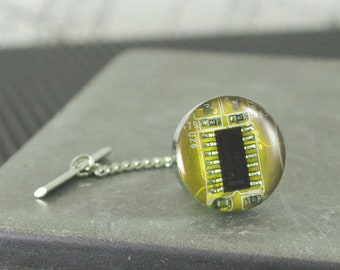 May include: A silver tie tack with a round, clear glass top. The glass top features a yellow circuit board with black components and the text "U26 79".