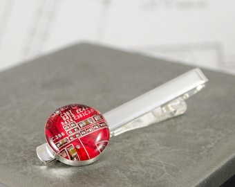 May include: A silver tie clip with a red circular glass cabochon featuring a circuit board design. The circuit board has text including "C386", "R473", and "EC42".