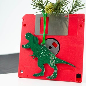 Könnte beinhalten: Eine rote Diskette mit einem grünen, aus einer Platine gefertigten Dinosaurier-Anhänger. Der Dinosaurier ist an einem grünen Band befestigt.