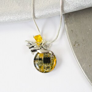 Könnte beinhalten: Eine silberne Kettenkette mit einem Anhänger, der einen kleinen silbernen Engel zeigt, der eine runde, gelbe und schwarze Platine hält. Die Platine ist in klarem Harz gegossen. Ein kleiner gelber Kristall hängt an der Hand des Engels.