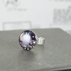 Puede incluir: Un anillo de plata con un diseño redondo de placa de circuito púrpura y dorado.