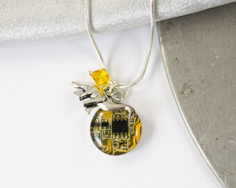 May include: A silver chain necklace with a pendant featuring a small silver angel holding a round, yellow and black circuit board. The circuit board is encased in clear resin. A small yellow crystal dangles from the angel's hand.