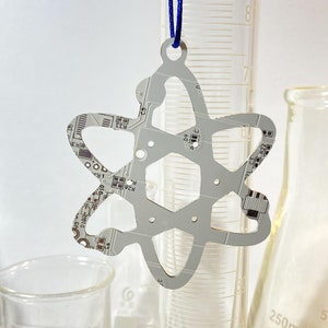 May include: A silver atom-shaped ornament with a circuit board design. The ornament is hanging from a blue string and is in front of a graduated cylinder and a beaker.