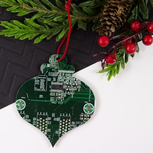 May include: Green circuit board Christmas ornament with a red string for hanging. The ornament has a unique teardrop shape and is decorated with electronic components. The background includes a sprig of evergreen, red berries, and a black and white patterned surface.