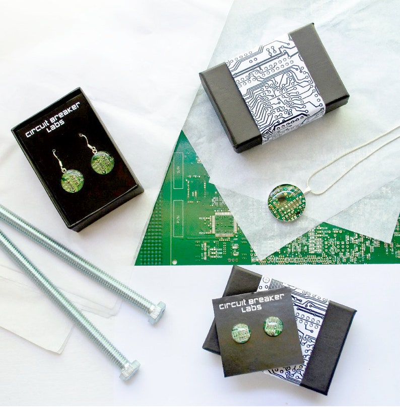 May include: Two black boxes with the text "Circuit Breaker Labs" on the front, each containing a pair of green and gold earrings. The earrings are made from circuit boards. The boxes are sitting on a white surface with a green circuit board in the background.