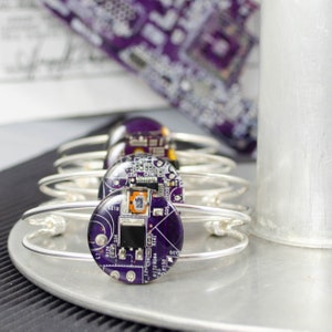 May include: A stack of silver-toned metal bangles, each featuring a circular, purple circuit board design. The circuit boards have various electronic components. The bangles are displayed on a silver surface, with a purple patterned fabric in the background.