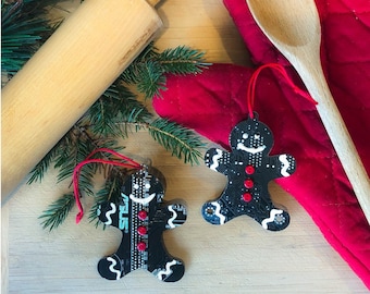 May include: Two black gingerbread man ornaments with red hanging strings. The ornaments have white accents and red button details. One ornament has the word "ASUS" printed on it. The ornaments are on a wooden surface with a rolling pin and greenery.