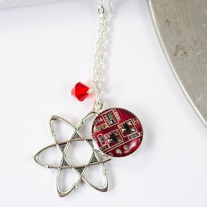 Könnte beinhalten: Eine silberne Halskette mit einem roten Kristall-Anhänger und einem Anhänger, der ein silbernes Atom-Symbol und ein rundes rotes Harzstück mit einem Leiterplatten-Design zeigt.