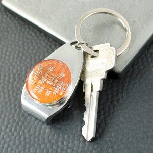 May include: A silver key ring with a round, orange, circuit board design. The keyring has a bottle opener attached to it. A silver key is attached to the keyring.