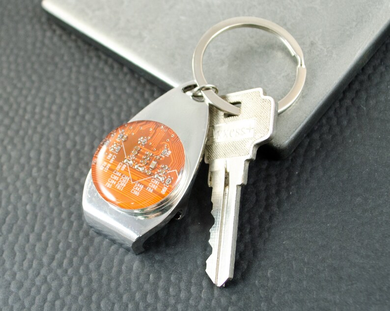 May include: A silver key ring with a round, orange, circuit board design. The keyring has a bottle opener attached to it. A silver key is attached to the keyring.