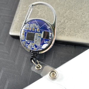 May include: A silver retractable badge holder with a blue and silver circuit board design. The holder has a swivel clip and a clear plastic strap.