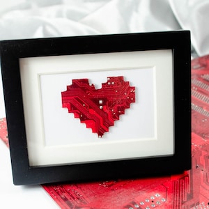 Könnte beinhalten: Eine rote Platine in Herzform ist in einem schwarzen Rahmen gerahmt. Der Rahmen liegt auf einer roten Platine.