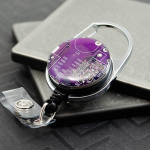 May include: A silver retractable badge reel with a purple circuit board design. The reel has a swivel clip and a clear plastic strap.