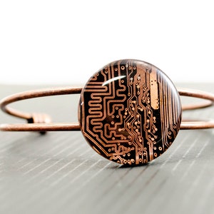 Pode incluir: Um bracelete de fio de cobre com um charme redondo de resina preta e cobre com um design de placa de circuito.