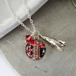 Könnte beinhalten: Eine silberne Kettenkette mit einem roten Leiterplattenanhänger, einem silbernen Raketenanhänger und einem roten Kristallanhänger.