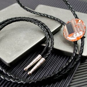 May include: A black braided bolo tie with a silver clasp and a round orange and silver circuit board pendant.