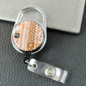 Puede incluir: Un portagafete retráctil plateado con un diseño redondo, transparente, naranja y blanco de placa de circuito. El portagafete tiene un clip de metal y una correa de plástico transparente.