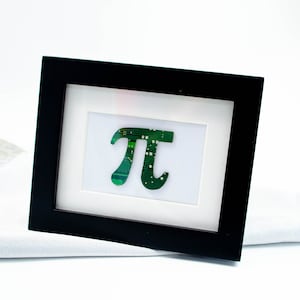 May include: A framed artwork displaying the Pi symbol, constructed from green circuit board material. The symbol is set on a white background within a black rectangular frame. The design has a modern, tech-inspired aesthetic.